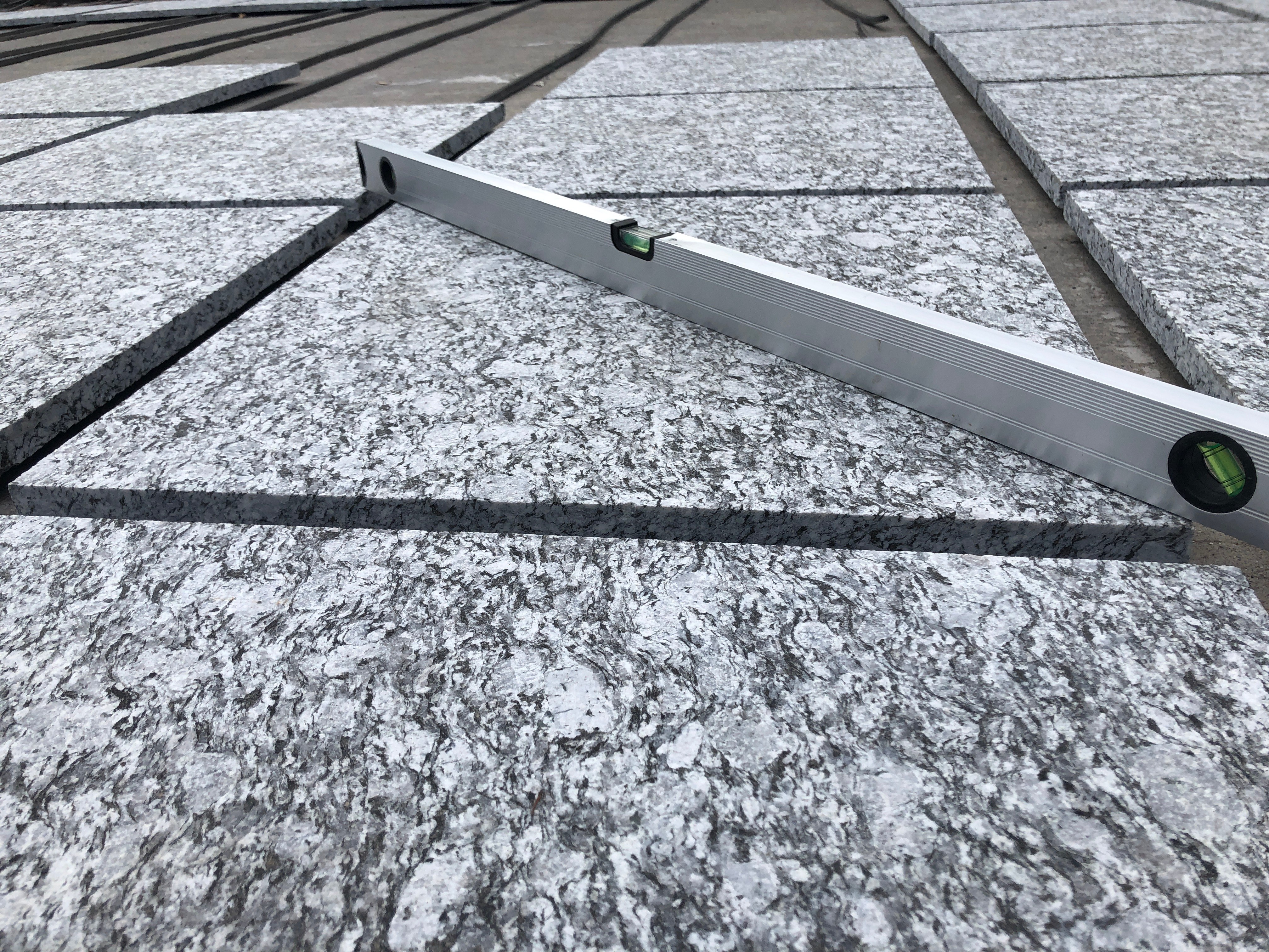 Aluminium spirit level laid diagonally across flamed sea-wave grey granite paving tiles to check surface flatness during dry-laying