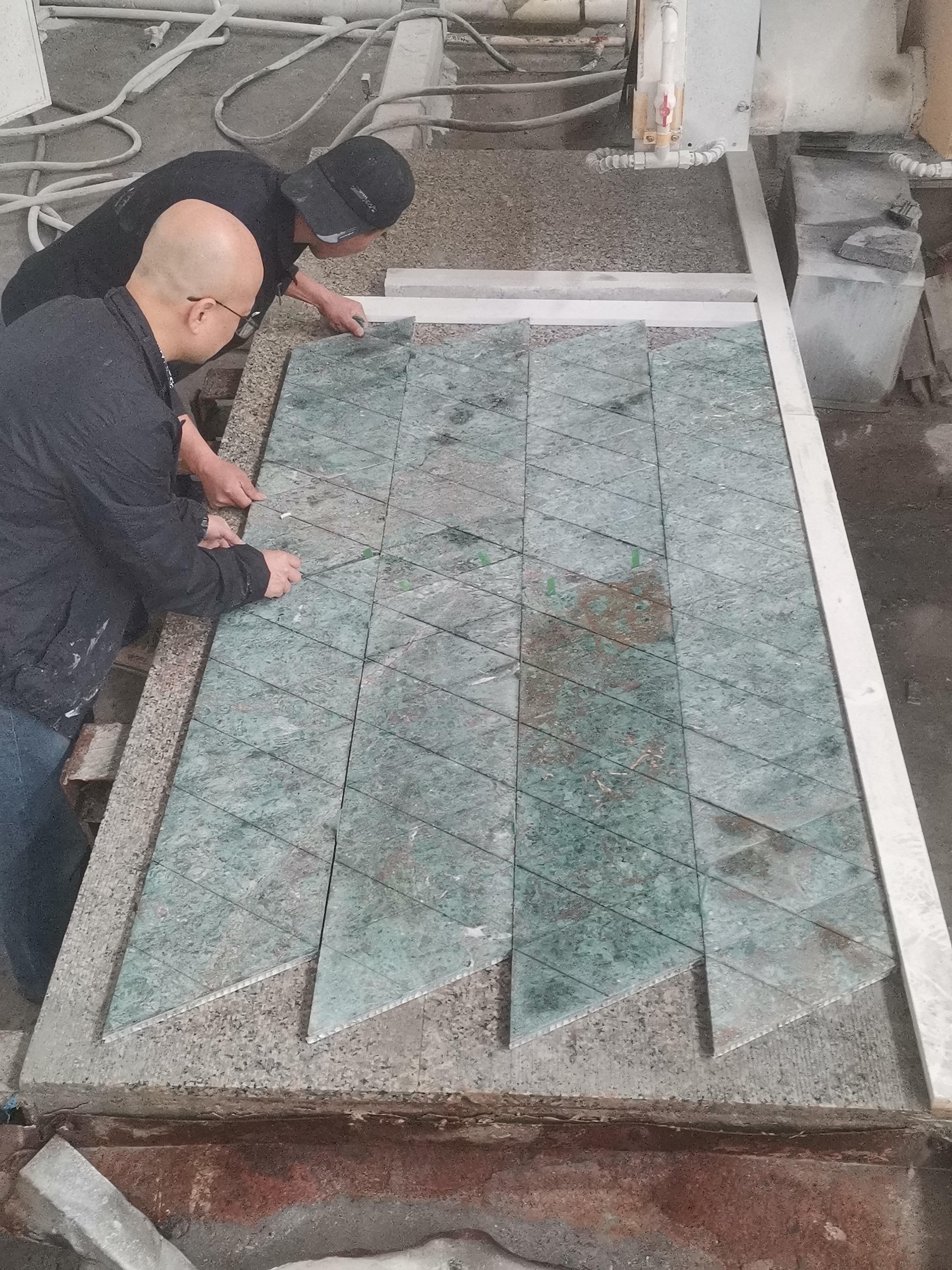 Two Sun Young Stone craftsmen dry-laying and seam-matching triangular fluted Rainforest Green marble tiles on a factory platform